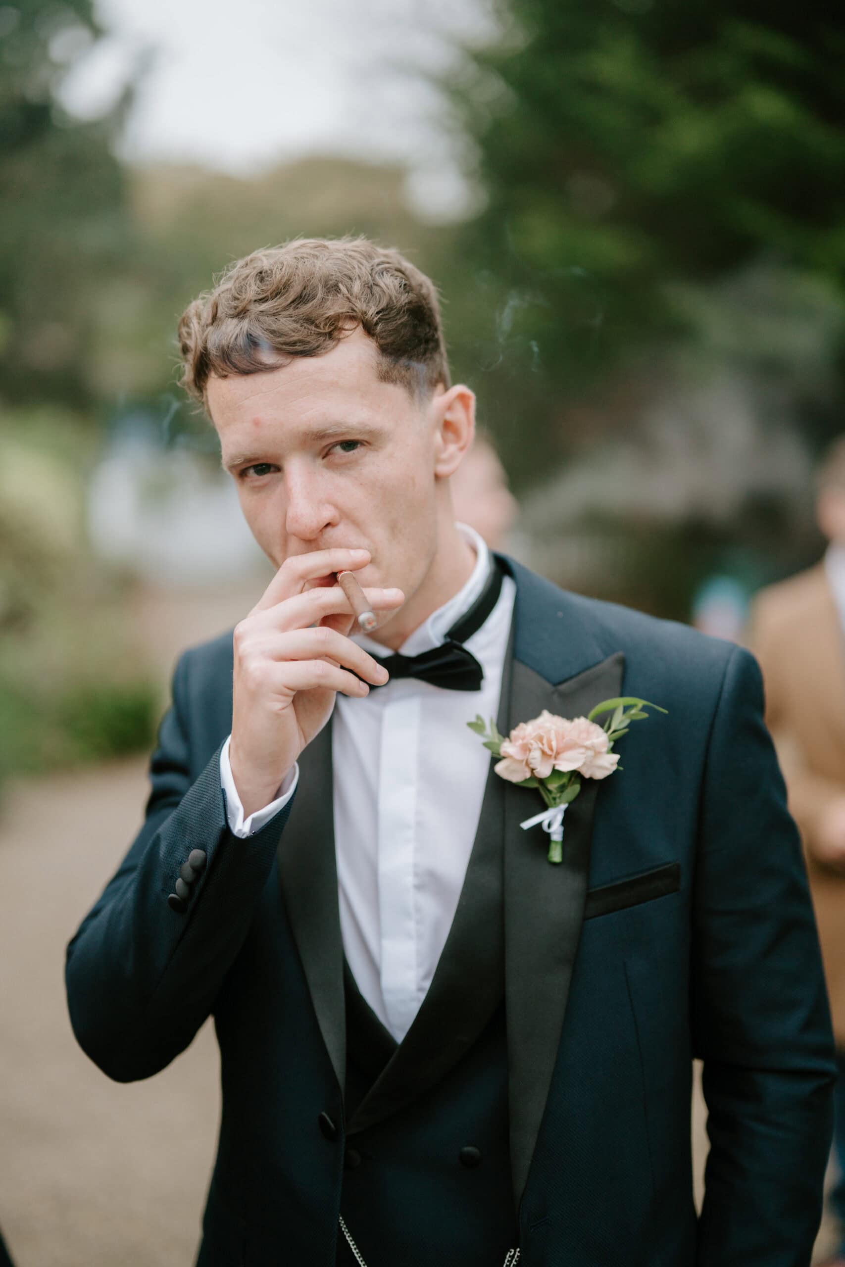 A man in a formal suit with a bow tie stands outdoors, smoking a cigarette. He wears a boutonniere with a pink flower, captured elegantly by the Kent wedding photographer. The background is blurred, with hints of foliage and another person in the distance. Image by Pearce Wedding Photography.