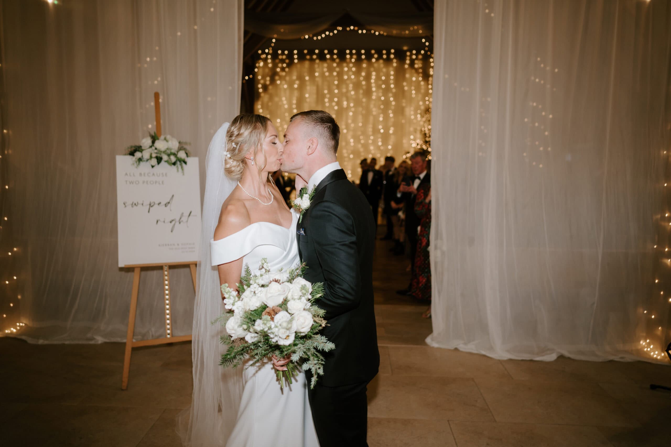 A bride and groom share a kiss at their wedding, captured by a talented Kent wedding photographer. Surrounded by twinkling lights and sheer curtains, the bride holds a bouquet of white flowers. A welcome sign is visible in the background as guests look on from behind. Image by Pearce Wedding Photography.