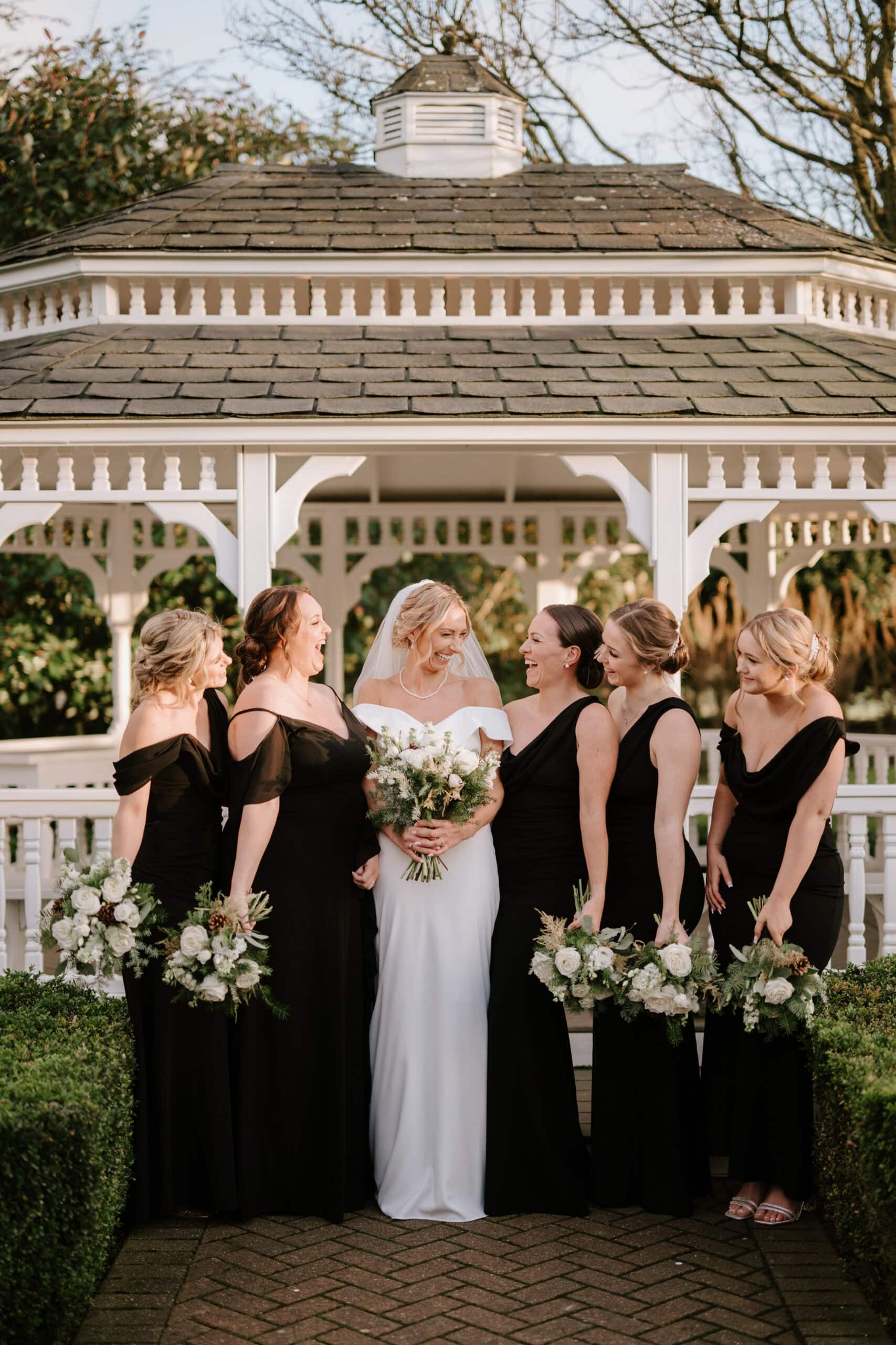 A bride in a white dress and veil stands with five bridesmaids in black dresses, all holding bouquets of flowers. They are smiling in front of the charming white gazebo at The Old Kent Barn, surrounded by lush greenery, capturing a perfect wedding moment. Image by Pearce Wedding Photography.