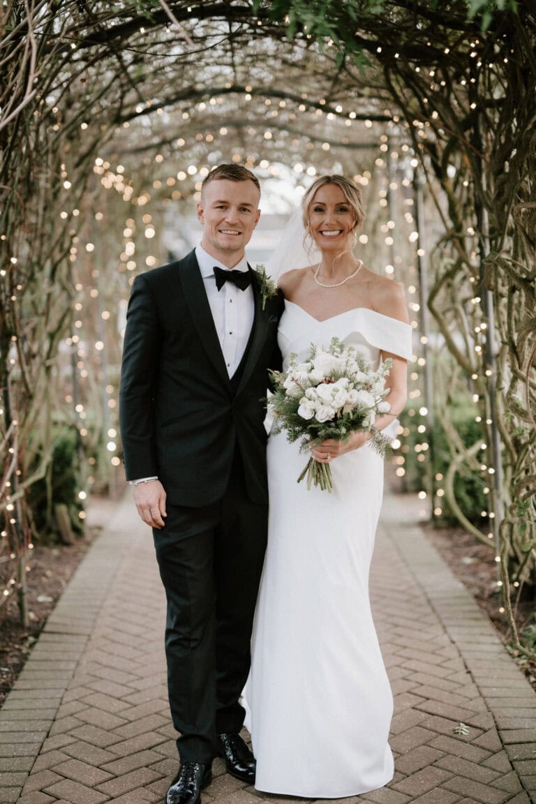 Under the vine-covered archway adorned with fairy lights at The Old Kent Barn, a bride and groom smile warmly at the camera. She holds a bouquet of white flowers in her off-shoulder gown, while he stands beside her in a sharp black tuxedo, encapsulating the perfect wedding moment. Image by Pearce Wedding Photography.
