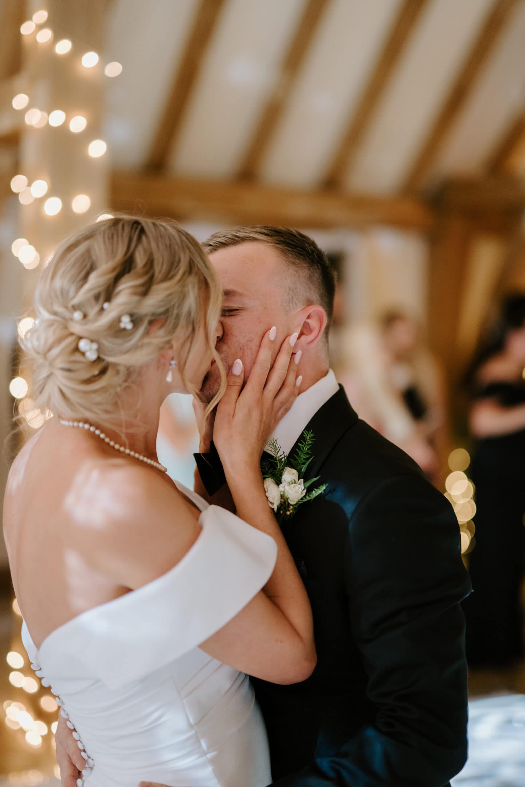 A bride and groom kiss during their wedding ceremony. The bride wears an off-shoulder white dress with a pearl hair accessory, and the groom is in a black suit with a boutonniere. Captured by a Kent wedding photographer, soft fairy lights create a warm and romantic atmosphere. Image by Pearce Wedding Photography.