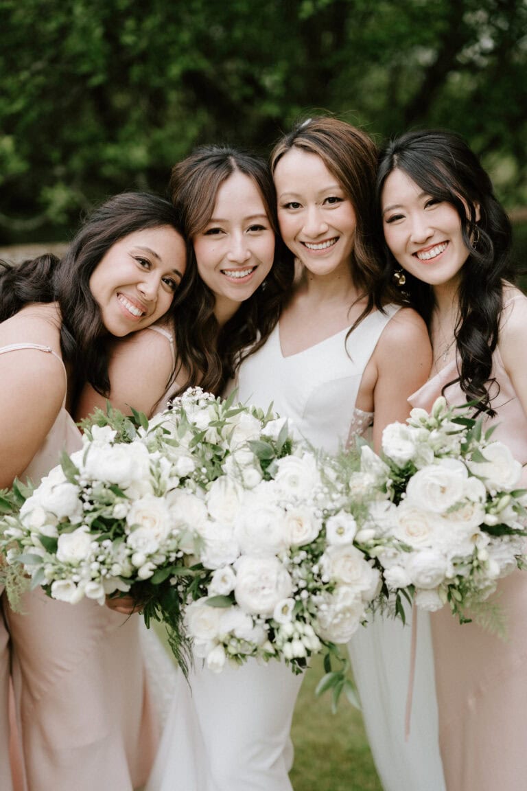Bridesmaids smiling holding white rose bouquets outdoors.