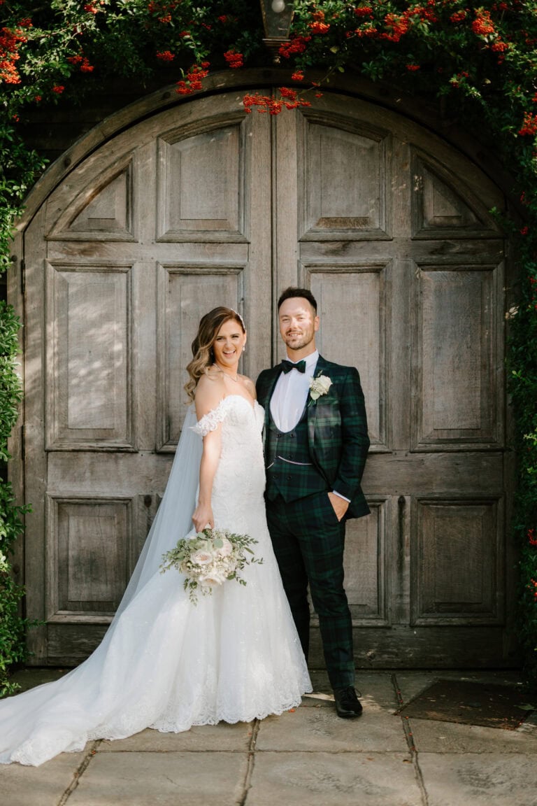 A bride and groom stand smiling in front of a large wooden door, captured perfectly in this exquisite wedding photography. The bride holds a bouquet while wearing a white gown, and the groom dons a plaid suit. Red flowers and green foliage beautifully frame the arch above them. Image by Pearce Wedding Photography.