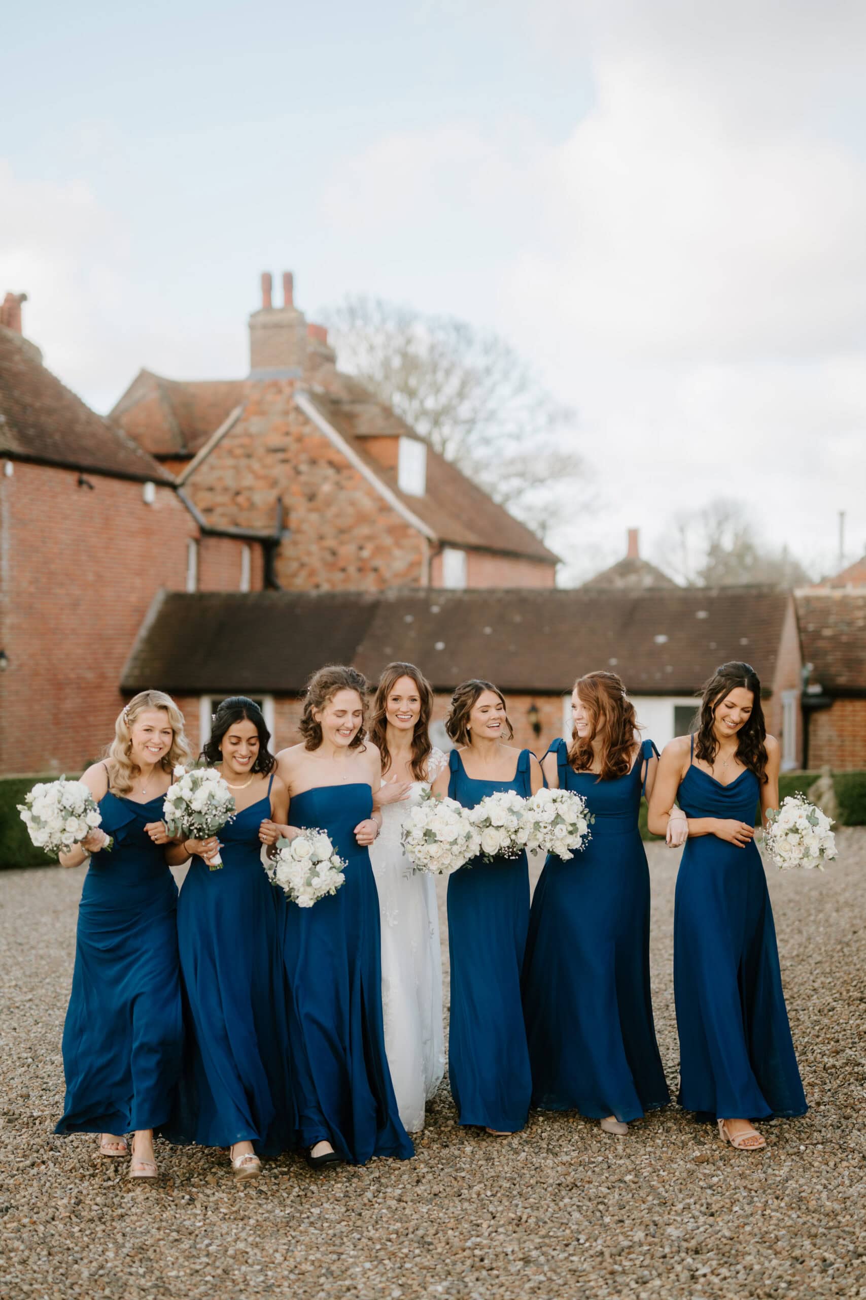 A bride stands with six bridesmaids, all in matching blue dresses, holding bouquets of white flowers. Captured by a talented Kent wedding photographer, they smile as they walk outdoors in front of brick buildings under a partly cloudy sky. Image by Pearce Wedding Photography.