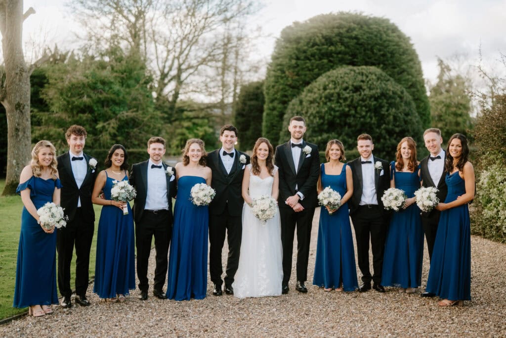 A wedding party poses outdoors on a gravel path at Winters Barns. The bride in a white gown stands with the groom in a black suit, surrounded by bridesmaids in blue dresses and groomsmen in black suits, all holding bouquets. Trees and bushes provide a charming backdrop. Image by Pearce Wedding Photography.