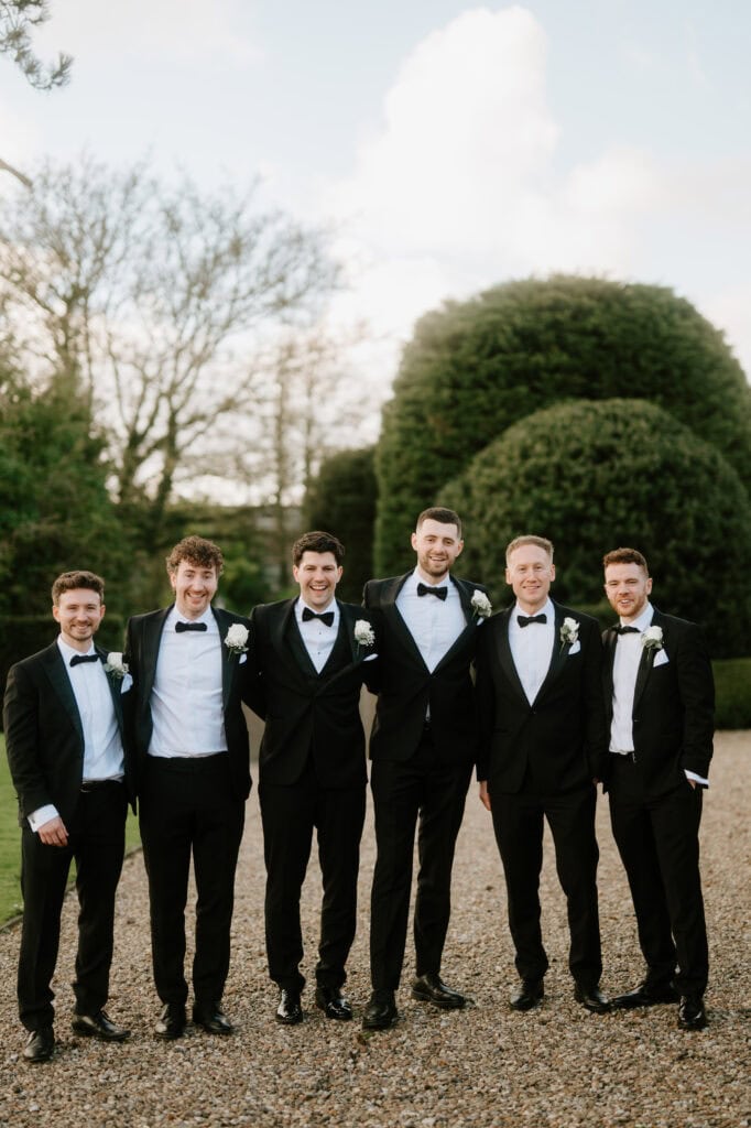 Six men in tuxedos stand smiling on a gravel path at Winters Barns. They have white boutonnières, surrounded by greenery that complements the wedding's vibrant spirit. The sky is clear, creating a bright and cheerful atmosphere. Image by Pearce Wedding Photography.
