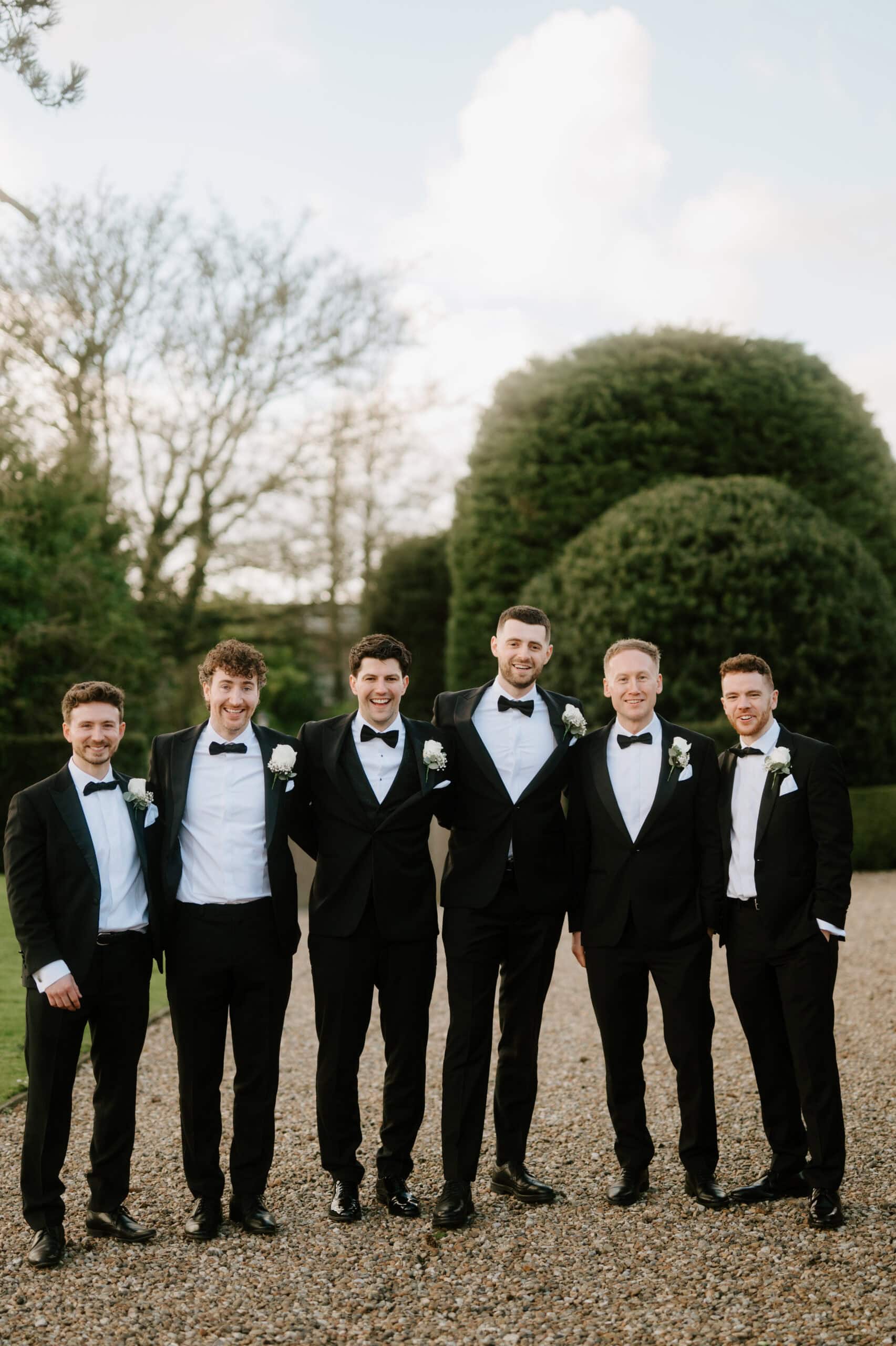 Six men in tuxedos stand smiling on a gravel path at Winters Barns. They have white boutonnières, surrounded by greenery that complements the wedding's vibrant spirit. The sky is clear, creating a bright and cheerful atmosphere. Image by Pearce Wedding Photography.