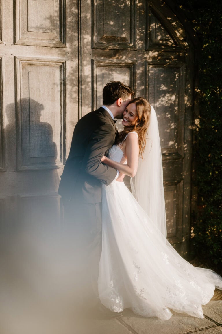At Winters Barns, a groom in a suit kisses his smiling bride on the cheek. The bride, radiant in her white wedding dress and veil, stands next to him as dappled sunlight dances through the wooden door behind them, casting playful shadows that echo their joyful wedding day. Image by Pearce Wedding Photography.