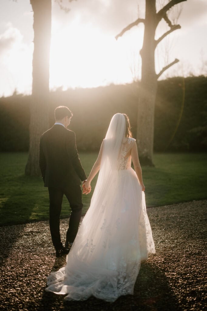 A bride and groom walk hand in hand on the gravel path of Winters Barns, lit by the warm glow of a setting sun. The bride wears a flowing white gown with a veil, and the groom is in a dark suit. Tall trees and a hedge form the serene wedding backdrop. Image by Pearce Wedding Photography.
