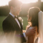 A couple in wedding attire embrace outdoors at sunset, framed by the picturesque charm of Winters Barns. The groom dons a dark suit and bow tie, while the bride wears a white gown and veil. Sunlight creates a warm glow and lens flare around them. Image by Pearce Wedding Photography.