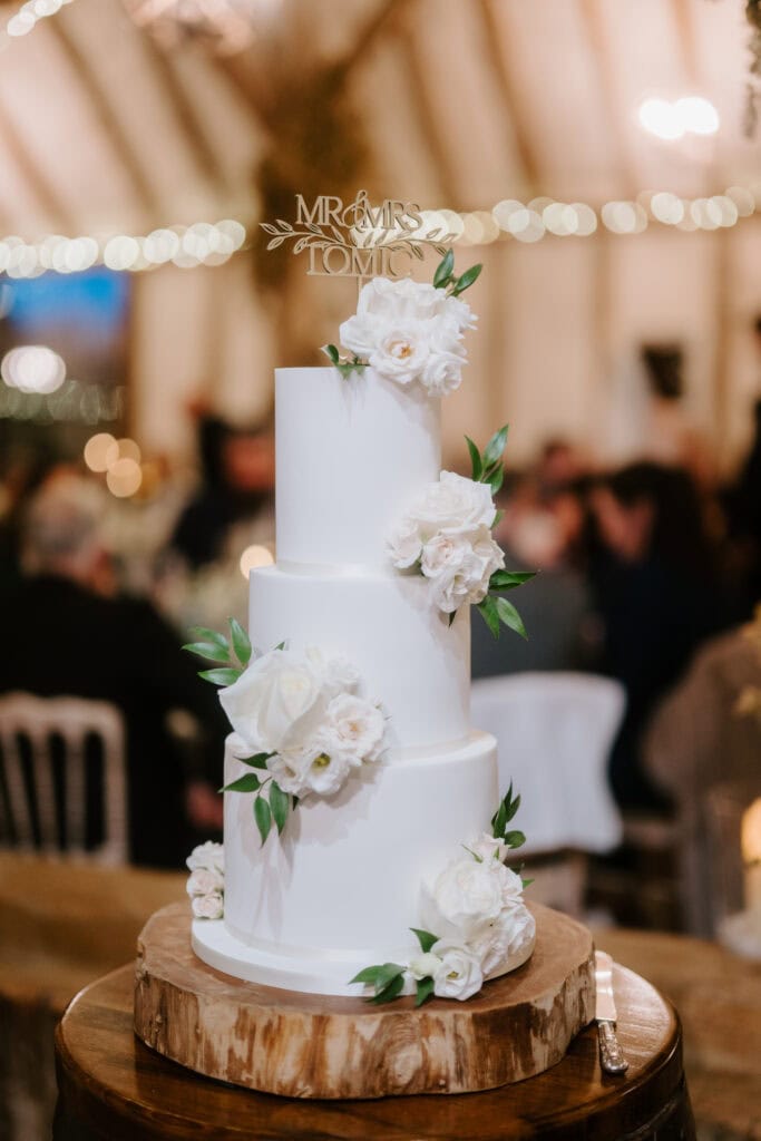 A three-tiered white wedding cake is adorned with white roses and green leaves, elegantly capturing the essence of a wedding at Winters Barns. It sits on a wooden base with small lights, blurred people in the background, and a "Mr & Mrs" topper crowning the scene. Image by Pearce Wedding Photography.