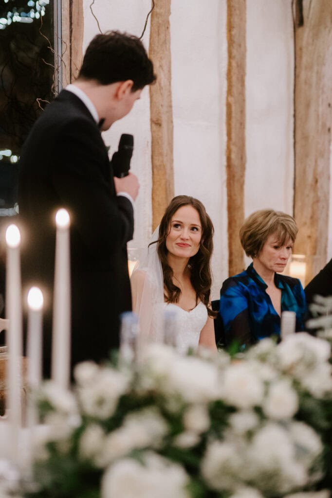 At a beautiful wedding reception, a man in a suit speaks into the microphone. The bride, radiant in her white gown, smiles up at him. Beside her, an older woman in blue gazes down thoughtfully. White flowers and candles shimmer on the table, capturing the romance of Winters Barns. Image by Pearce Wedding Photography.