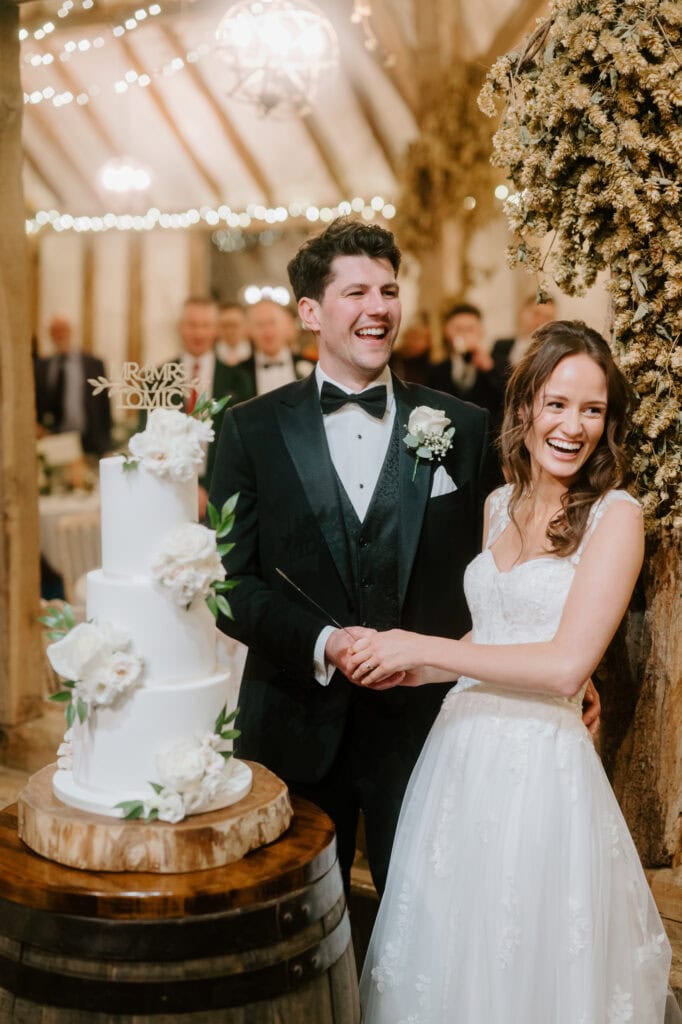 In the cozy charm of Winters Barns, a bride and groom joyfully cut a white tiered wedding cake adorned with flowers. Bathed in warm lighting, surrounded by guests in a rustic venue, the groom wears a tuxedo while the bride dazzles in her white gown. Image by Pearce Wedding Photography.