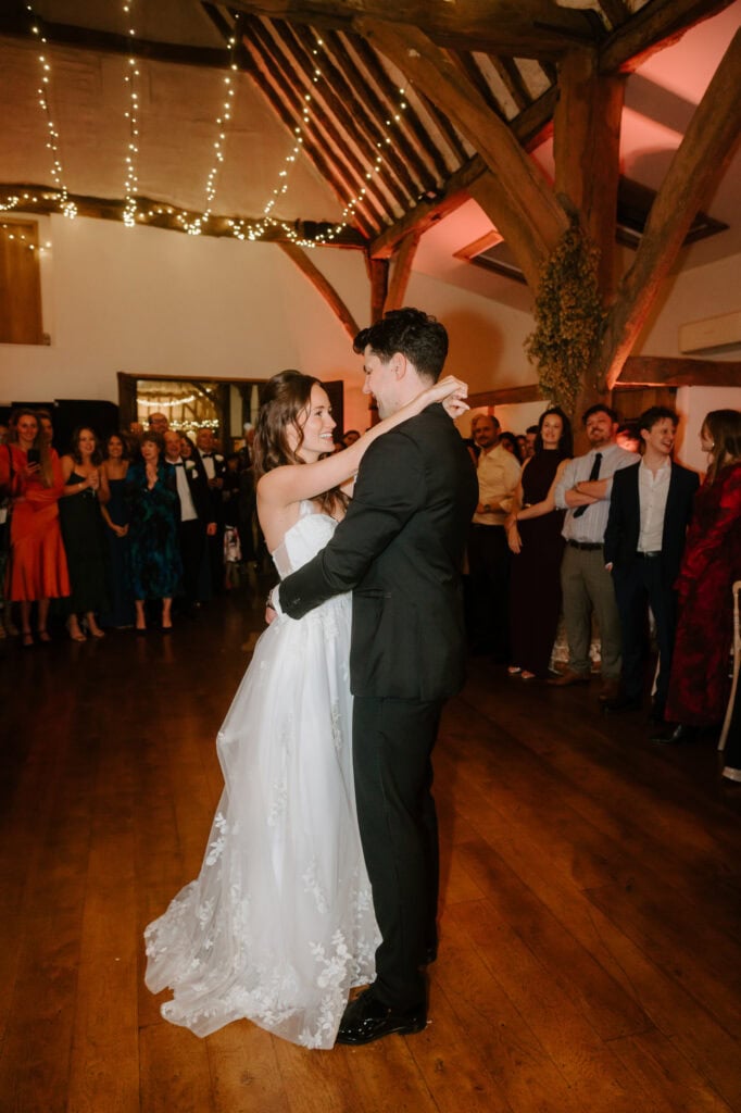 A couple dances closely on a wooden floor in a warmly lit room. The woman wears a white gown, and the man is in a dark suit. Guests watch them, standing around the edges of the wedding venue under a ceiling decorated with string lights. Image by Pearce Wedding Photography.