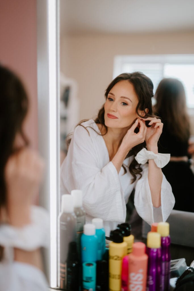 A woman in a white robe adjusts her earring while looking in a mirror, various hair products lined up on the counter. Her long, wavy hair cascades down as she wears a content expression, capturing a serene wedding preparation moment. Image by Pearce Wedding Photography.