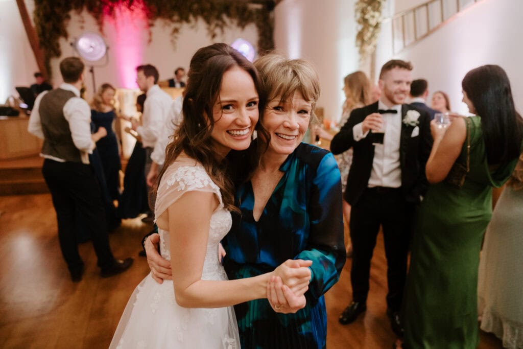 At Winters Barns, a bride in a white dress smiles and embraces an older woman in a blue dress on the dance floor. Surrounded by guests dancing and chatting, the scene is illuminated by soft lighting with enchanting greenery decor in the background, making for an unforgettable wedding. Image by Pearce Wedding Photography.
