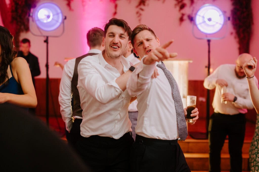 Two men in dress shirts and ties enthusiastically pose and gesture toward the camera at a lively wedding event. They stand close together against a vibrant backdrop at Winters Barns, with pink lighting and decorative elements. Other attendees appear blurred in the background. Image by Pearce Wedding Photography.