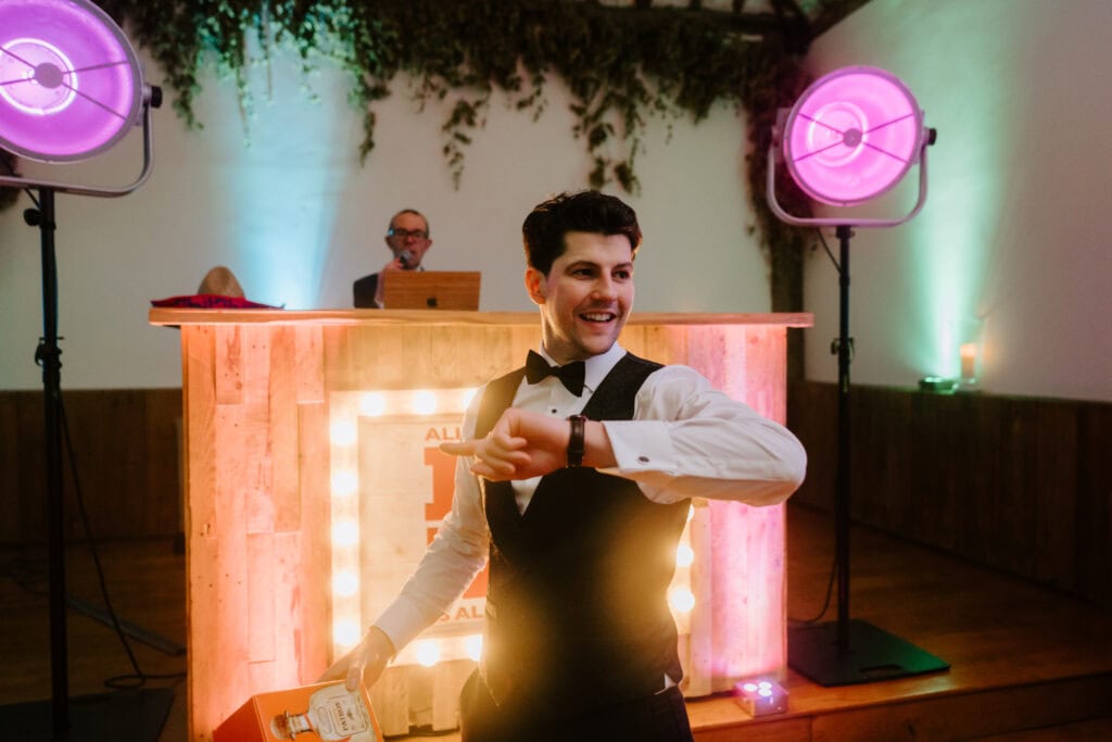 A person in formal attire, wearing a bow tie and vest, smiles and gestures with one arm in front of a wooden bar lit with marquee lights at a Winters Barns wedding. A DJ is in the background behind the bar, colorful lights illuminating the celebratory scene. Image by Pearce Wedding Photography.