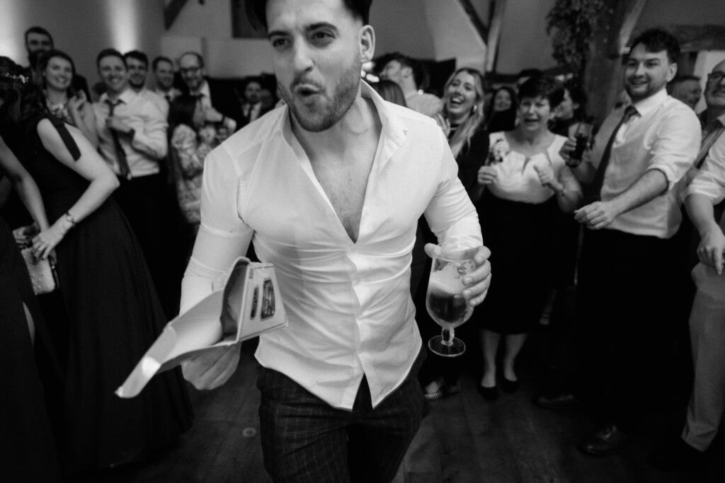 In a black and white photo, a man in a white shirt enthusiastically dances at Winters Barns with a drink in one hand and wedding papers in the other, surrounded by a cheering crowd. The atmosphere is festive and energetic. Image by Pearce Wedding Photography.