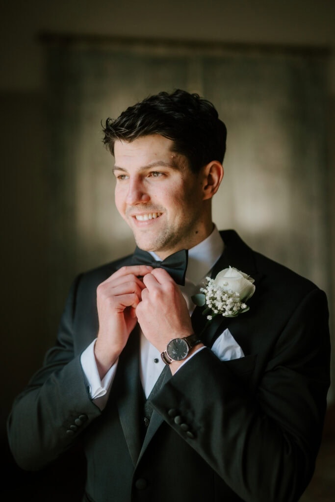 A man in a dark suit adjusts his bow tie, smiling. Dressed for the Winters Barns wedding, he wears a white shirt, a watch, and has a boutonniere with a white rose and baby's breath on his lapel. The background is softly blurred. Image by Pearce Wedding Photography.