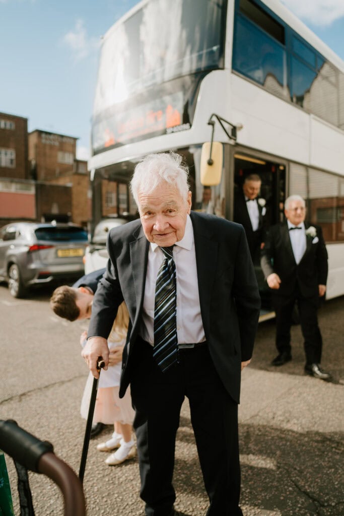 An elderly man with white hair, donning a suit and tie, stands in front of a white double-decker bus at Winters Barns, supporting himself with a walking stick. A child and two men, all dressed formally for the wedding, walk nearby. Image by Pearce Wedding Photography.