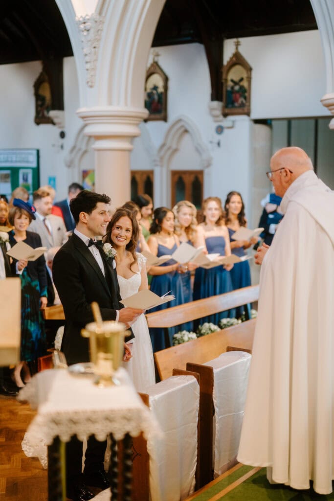 A couple stands at the altar, beaming during their wedding ceremony in a charming church. The groom dons a black tuxedo, while the bride dazzles in a white gown. As the priest speaks, the bridal party in blue dresses enhances this picturesque scene reminiscent of Winters Barns elegance. Image by Pearce Wedding Photography.