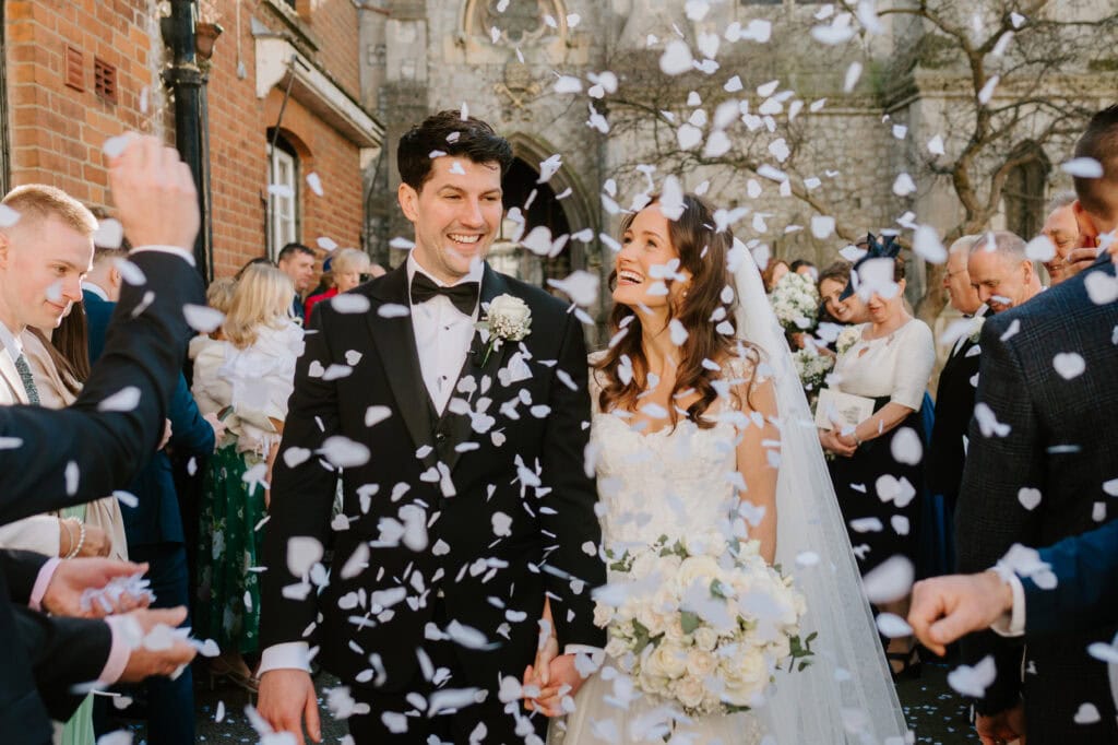 The image captures a joyful bride and groom holding hands, walking through a confetti shower outside Winters Barns. Surrounded by delighted guests, the bride showcases a bouquet of white flowers, epitomizing a perfect wedding day. Image by Pearce Wedding Photography.