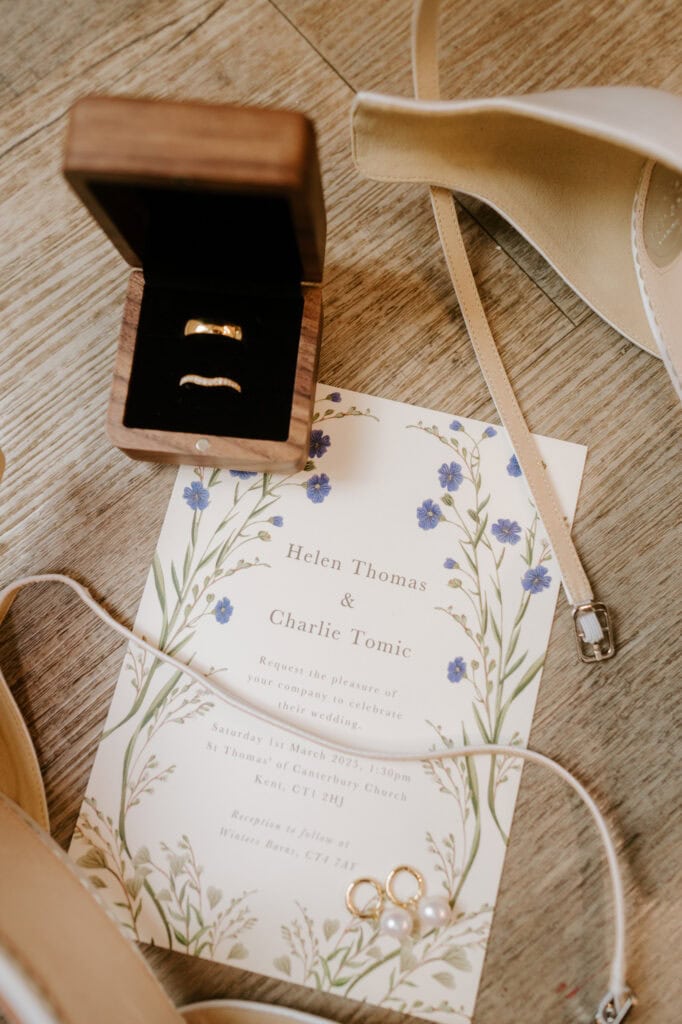 Wedding invitation with floral design for Helen Thomas and Charlie Tomic beside a wooden ring box with two rings. Nearby are beige high heels and a pearl bracelet. The invitation details a March 11, 2023 wedding at St Thomas' Church in Kent, CT1 7NU, followed by a reception at Winters Barns. Image by Pearce Wedding Photography.