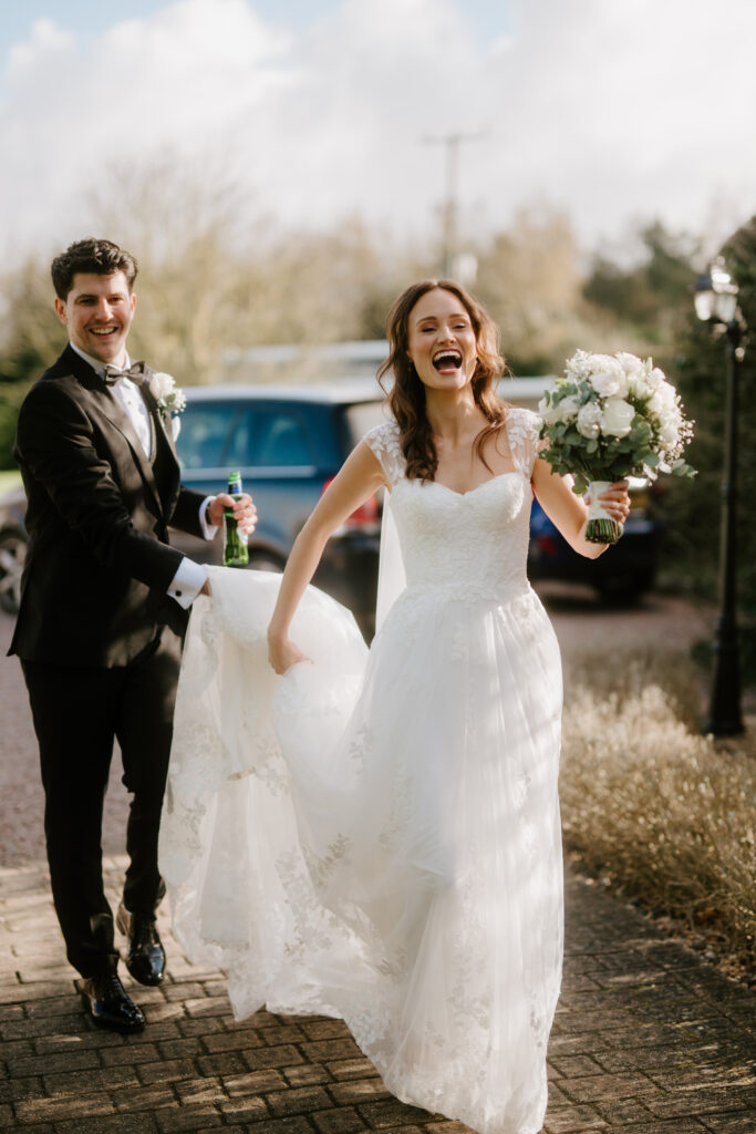 The bride and groom walk outside Winters Barns, smiling and laughing. The groom, in a black suit, holds a green bottle and the bride's train. The bride, in her white wedding gown, carries a bouquet of white flowers. A car and trees frame this charming scene. Image by Pearce Wedding Photography.