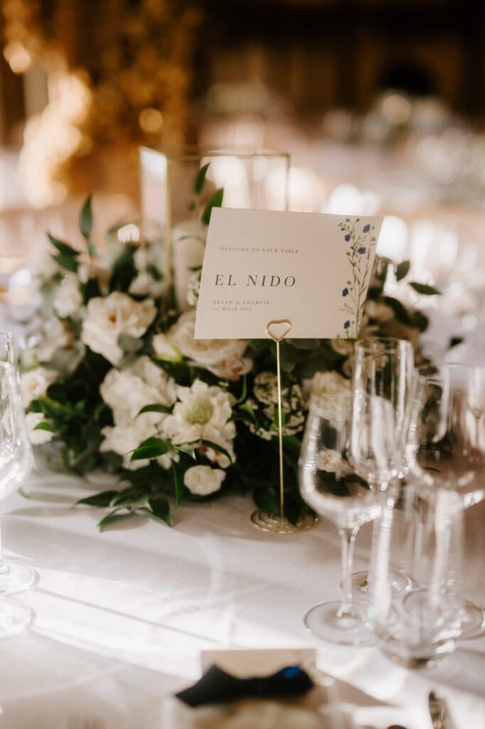 A beautiful wedding table centerpiece with white flowers and greenery graces the sunlit setting. Nestled among the blooms, a card labeled "El Nido" sits in a gold holder, while crystal wine glasses encircle the arrangement—a touch of elegance reminiscent of Winters Barns romance. Image by Pearce Wedding Photography.