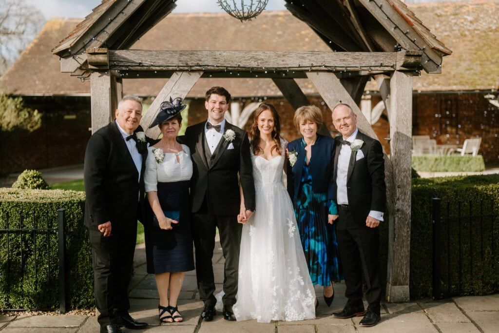 Bride and groom stand under a wooden arch at Winters Barns, flanked by two smiling women and two men, all dressed in formal wedding attire. The women wear elegant dresses and hats, while the men are in tuxedos. A rustic building is visible in the background. Image by Pearce Wedding Photography.