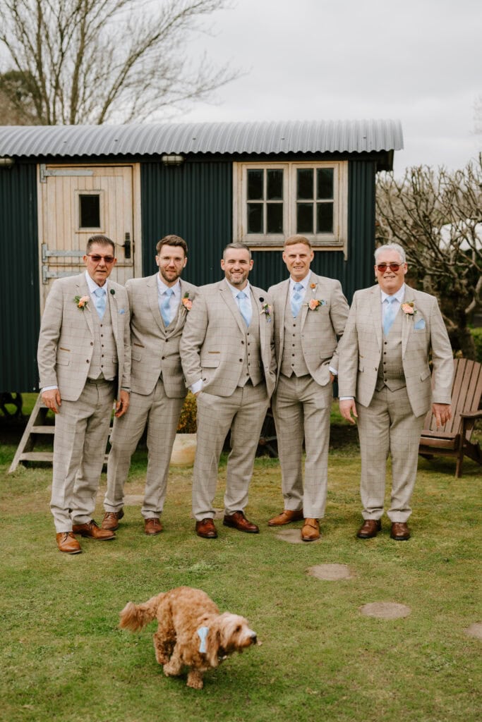 Groomsmen in suits with dog outside cottage.
