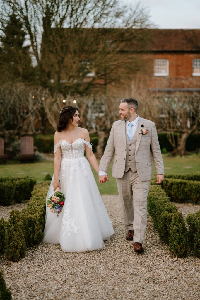A bride in a white gown holds a colorful bouquet as she walks hand in hand with a groom in a light gray suit. They stroll down a gravel path in the enchanting garden of Kent, reminiscent of The Secret Garden, with trimmed hedges and a brick building behind them. Image by Pearce Wedding Photography.