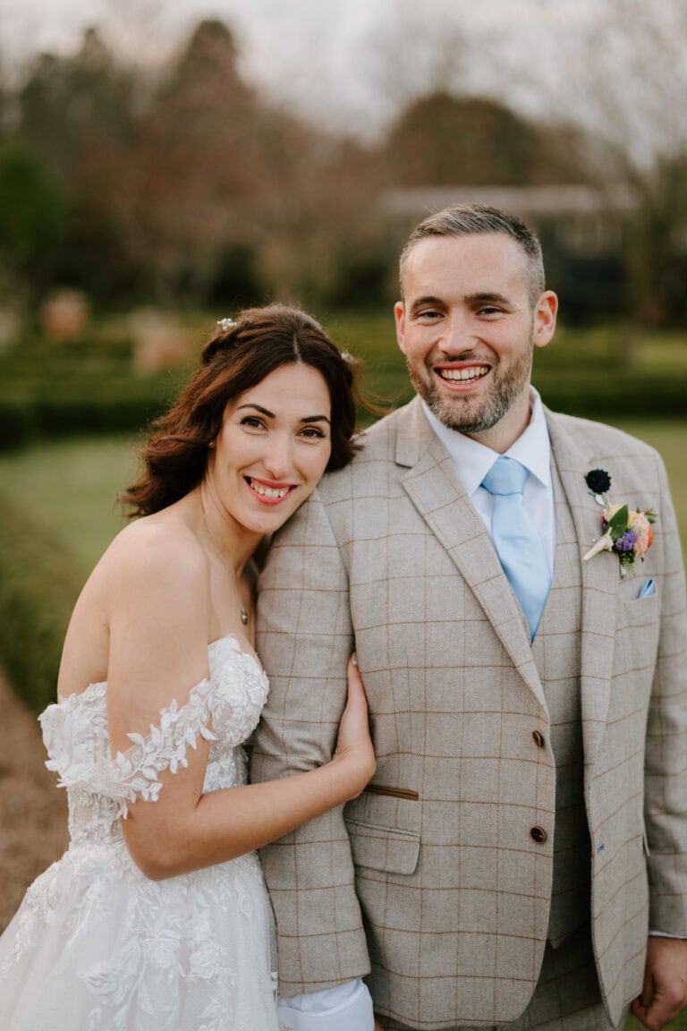A smiling couple poses outdoors on their wedding day at The Secret Garden in Kent. The bride, in an off-shoulder white lace gown, leans against the groom in a plaid suit with a light blue tie, surrounded by lush garden beauty. Image by Pearce Wedding Photography.