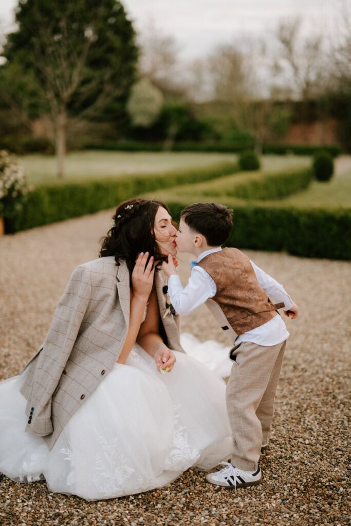 A bride in a white dress kneels on a gravel path, smiling as a young boy in a vest and bow tie kisses her cheek. The boy wears a matching tan outfit, including a checkered jacket draped over the bride's shoulders. A garden reminiscent of The Secret Garden blooms in the background. Image by Pearce Wedding Photography.