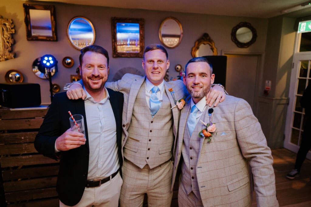 Three men in formal attire stand closely, smiling at the camera. Two wear matching light-colored suits, while the third opts for a dark jacket. In a room softly illuminated with framed mirrors, they appear ready for a joyous wedding celebration in Kent. One holds a drink, hinting at festivities to come. Image by Pearce Wedding Photography.