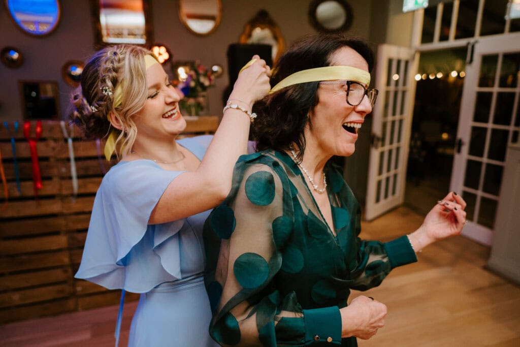 Two women are laughing as one ties a yellow ribbon over the other's eyes in a playful manner, reminiscent of a joyful scene from The Secret Garden. They are indoors, surrounded by wooden decor and mirrors. One wears a blue dress, while the other dons a green polka-dot dress. Image by Pearce Wedding Photography.