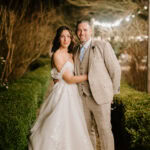 A couple stands beaming in a garden reminiscent of the Secret Garden. The bride, in an off-the-shoulder gown, and her groom, in a light plaid suit, embrace under twinkling string lights as manicured bushes line their enchanted Kent wedding pathway. Image by Pearce Wedding Photography.
