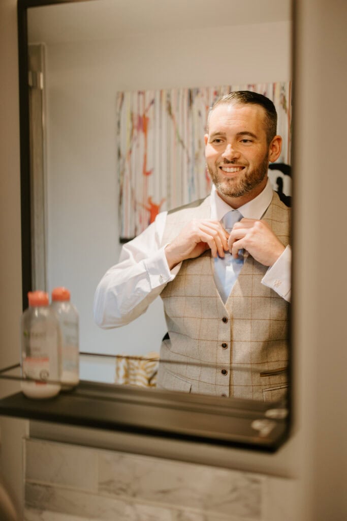 A man smiles as he stands in front of a mirror, adjusting his bow tie for the wedding. He is wearing a plaid vest over a white shirt. There are bottles on the shelf below the mirror, and a colorful abstract painting is reflected in the background, adding charm to this Kent setting. Image by Pearce Wedding Photography.