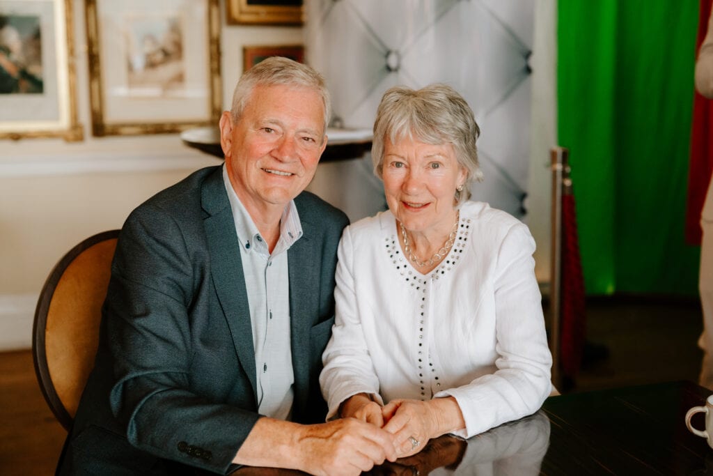 Elderly couple smiling and holding hands indoors.