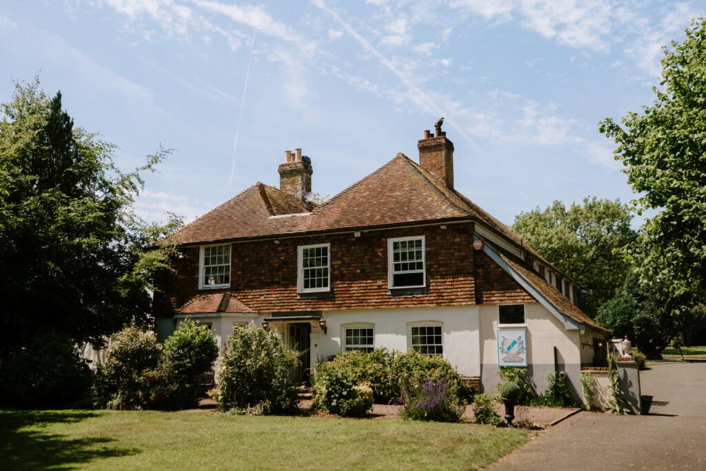 A traditional two-story house, reminiscent of Great Everden Manor, with a red-tiled roof, white lower walls, and brown upper brickwork, is surrounded by green trees and bushes under a partly cloudy blue sky—an idyllic wedding setting. Image by Pearce Wedding Photography.