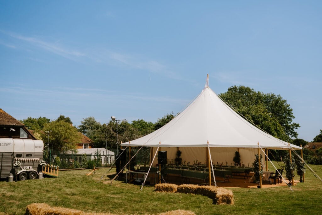 A large white event tent is set up outdoors on a grassy field at Great Everden Manor, surrounded by hay bales. Tables and chairs are arranged inside the tent, perfect for a charming wedding under a clear blue sky with trees and a building in the background. Image by Pearce Wedding Photography.