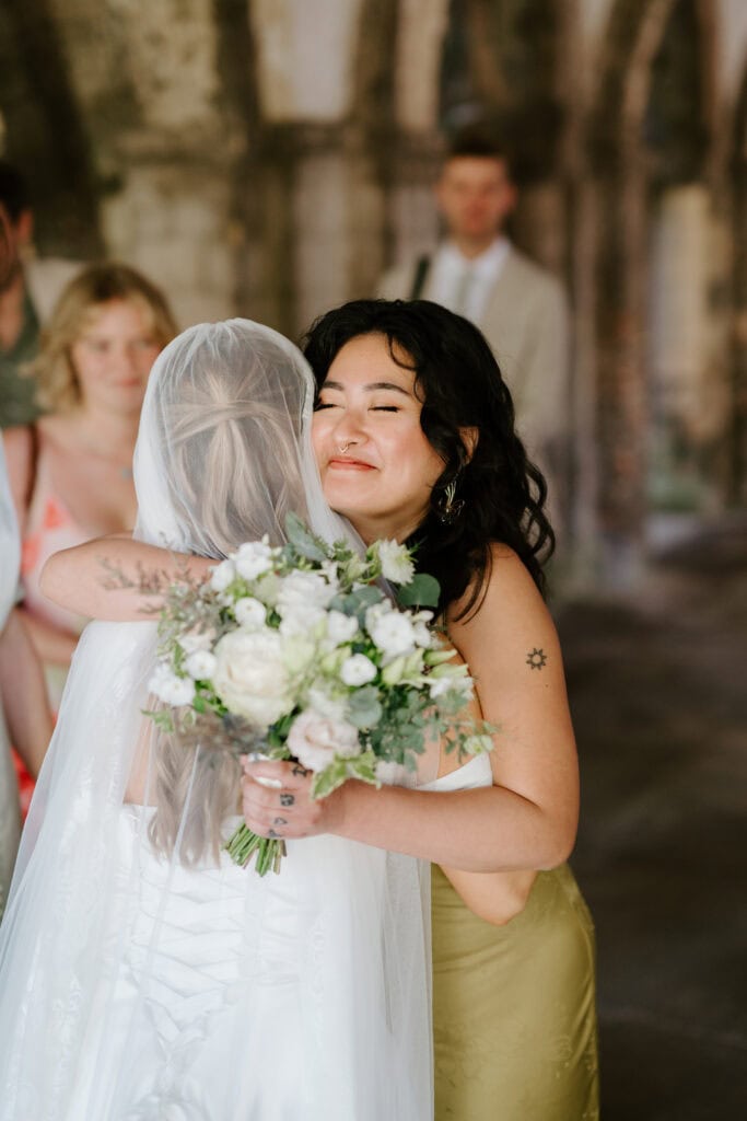 Bride hugging friend with bouquet indoors.