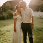 A woman in a white off-shoulder dress smiles as a man in a light shirt and dark pants kisses her on the head. They stand together on sunlit grass at Great Everden Manor, capturing a joyful wedding moment with buildings in the background. Image by Pearce Wedding Photography.