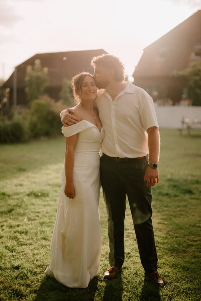 A woman in a white off-shoulder dress smiles as a man in a light shirt and dark pants kisses her on the head. They stand together on sunlit grass at Great Everden Manor, capturing a joyful wedding moment with buildings in the background. Image by Pearce Wedding Photography.