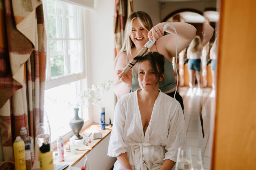 A woman in a white robe sits by a window at Great Everden Manor as another woman styles her hair with a curling iron—both smiling in a bright room filled with beauty products, flowers, and the excitement of a wedding day. Image by Pearce Wedding Photography.