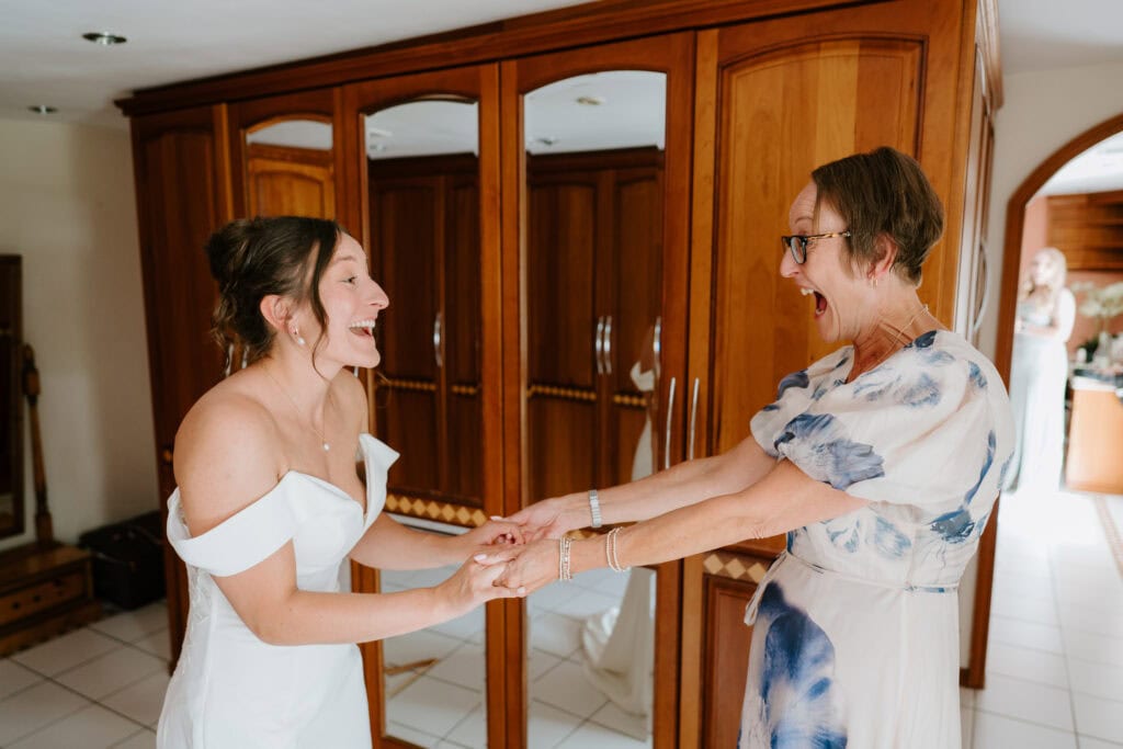 A bride in a white off-the-shoulder dress holds hands with an older woman in a floral dress. Both are smiling and laughing with excitement at their wedding in a sunlit room with wooden cabinets at Great Everden Manor. Image by Pearce Wedding Photography.