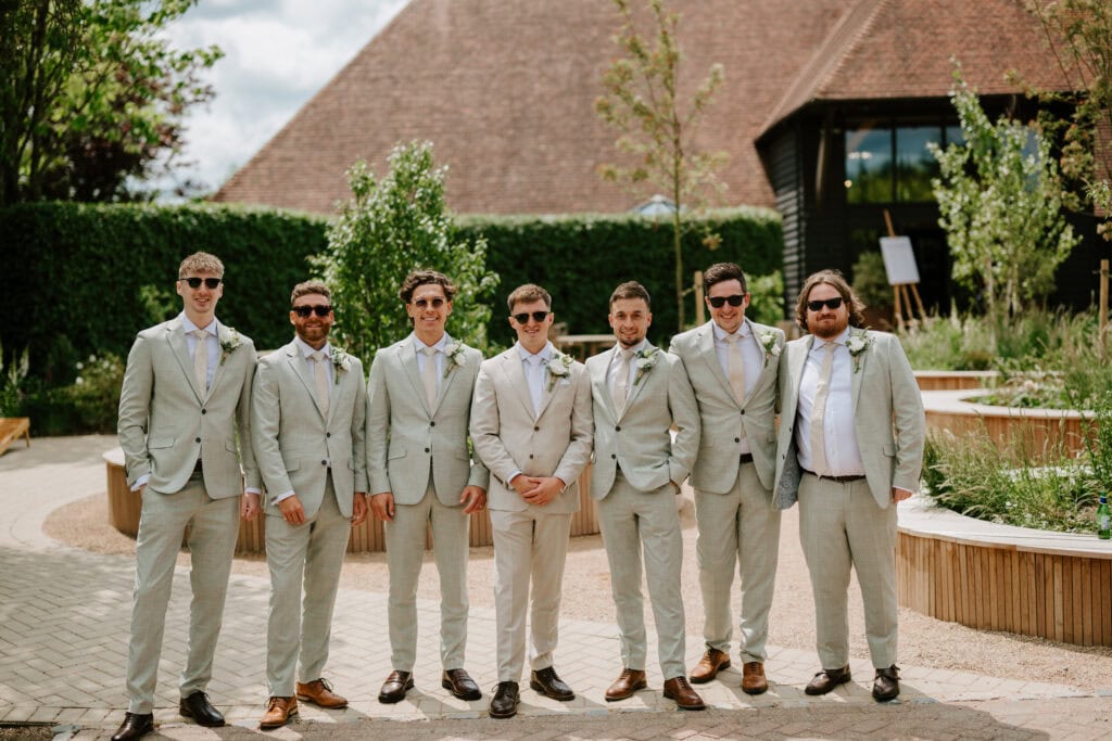 Seven groomsmen in suits outdoors, sunny garden setting.