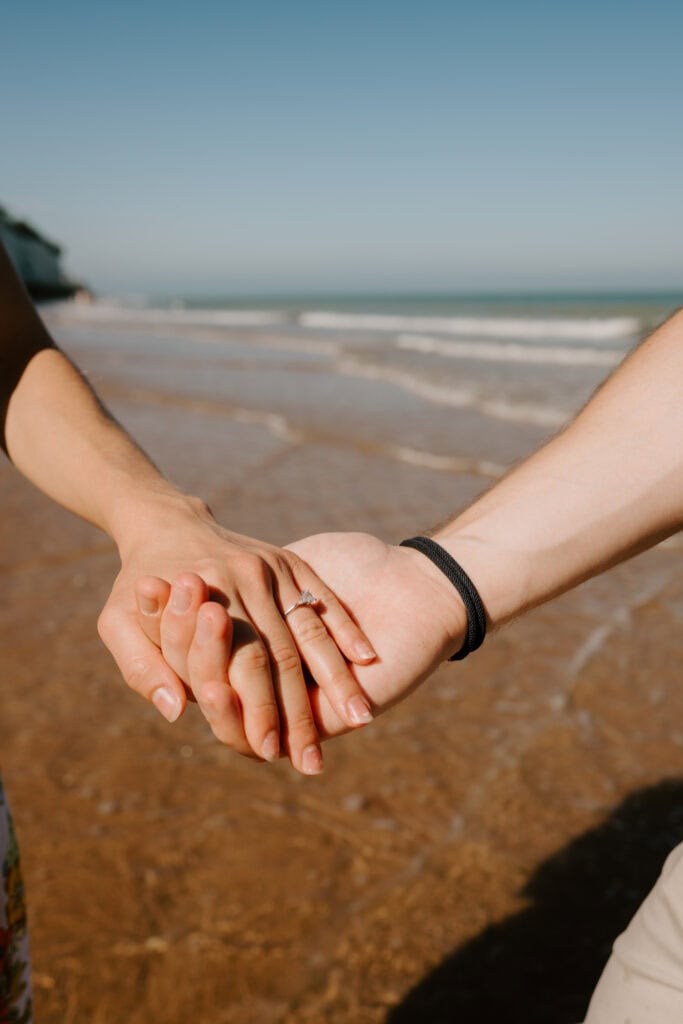 Couple holding hands on beach, engagement ring visible.