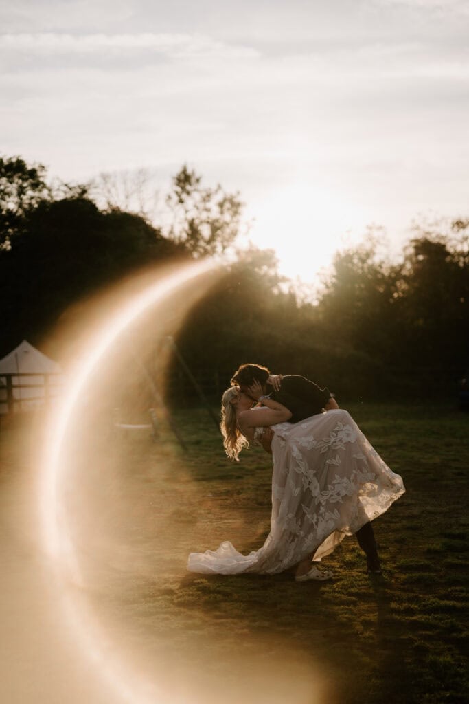 Couple kissing outdoors at sunset, lens flare visible.